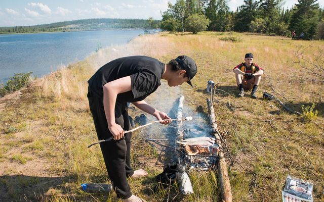 first nation teenage boy roasts marshmallow over fire next to a lake in thaidene nene