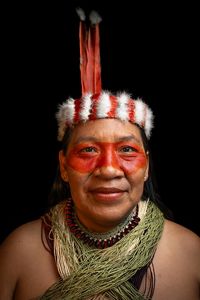 Retrato de mujer indígena de mediana edad, con pintura roja en la zona de sus ojos y pómulos, collar de fibras naturales en tonos verde y crema, collar de cuentas, cintillo de plumas y el pelo oscuro suelto hacia atrás. 