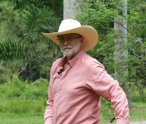 Un hombre con sombrero está de pie en un campo verde, mirando de frente a la cámara.