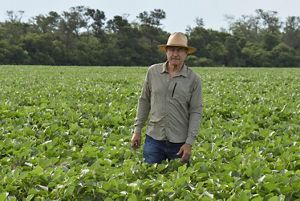  Un hombre con sombrero y camisa se encuentra de pie frente a un fondo de frondosos árboles verdes.