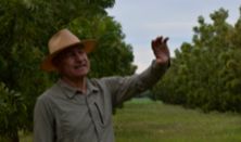  A man wearing a hat and shirt stands in front of a backdrop of lush green trees.