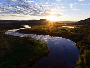 The sun rises over the Colorado River and the clouds and blue sky are reflected in the water.