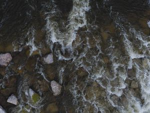 Aerial view features river water running over rocks with dark lighting.