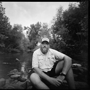 Black-and-white image of Jason Throneberry sitting at the edge of a river.