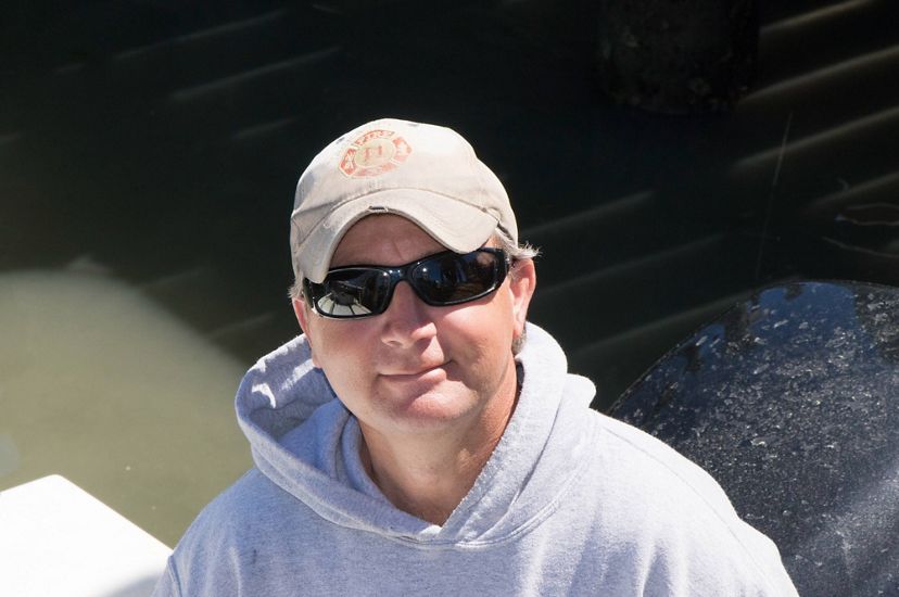 A man looking up at the camera from the deck of a small jonboat. He is wearing a gray hoodie and sunglasses. The sun reflects on the still water behind him.