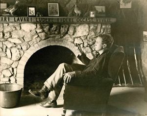 Author and historian John Joseph Mathews, seated in front of his fireplace with a pipe in hand. 