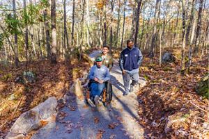 A man and his friend push another man using a wheelchair down a flat, winding path in the woods.
