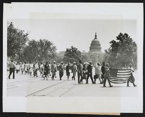 A group of Black Americans march together along an open plaza. Two men leading the group are in uniform and carry an American flag. The dome of the US Capitol building rises in the background.