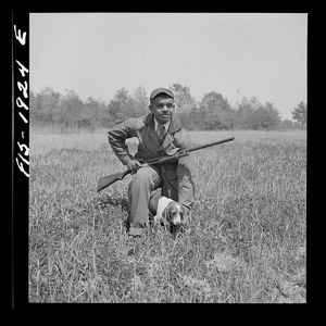 A man wearing a coat and tie kneels in an open field. A rifle rests on his knee and a small hunting dog crouches between the man's knees.