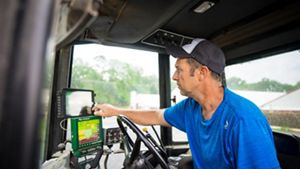 A man sits in the cab of a large tractor using small computer monitors to held determine the amount of fertilizer to apply to his field.