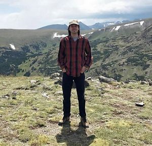 Joe Galarraga stands in a rocky clearing with tall mountains rising behind him.
