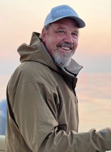 Geoff Smith, Marine Program Manager for The Nature Conservancy in Maine, smiles while fishing on the water, wearing a gray cap and brown jacket.