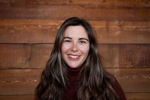 Head-and-shoulders portrait of a person with long hair and a dark sweater, photographed against a warm-toned wooden plank background.