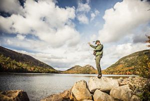 A park ranger holding a tool stands on a rock outcrop overlooking a lake. 
