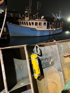 At a harbor at night, a weathered metal trap fills the foreground. Attached to the trap is a bright yellow cylindrical sensor secured with metal bands. A metal identification plate on the trap shows the markings 01033 and WFS125. In the background, a blue fishing vessel named Emma Rae is tied to the dock, reflected in the dark, still water under artificial lights.