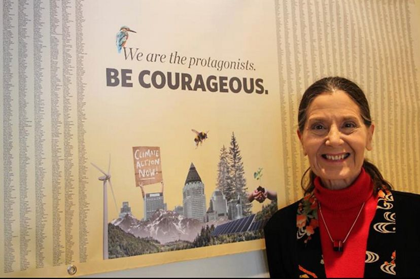 Lynn Scarlett stands in front of a poster that says, 'We are the protagonists. Be courageous', illustrated with buildings and nature and 25,000 signatures for climate action.