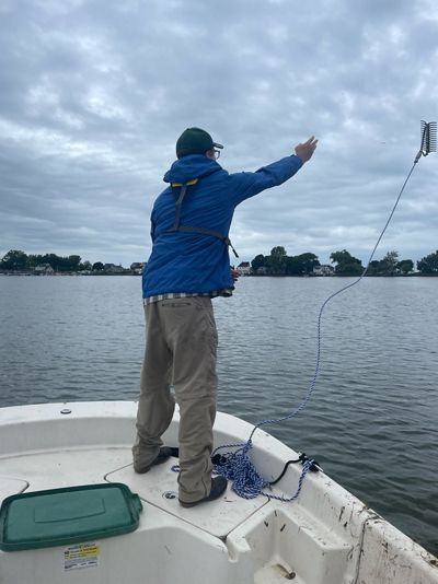 Gust Annis stands at the edge of a boat on Lake Ontario. Tethered to the boat is a rake that was thrown over the side. The water is still and the sky covered in dark clouds. 