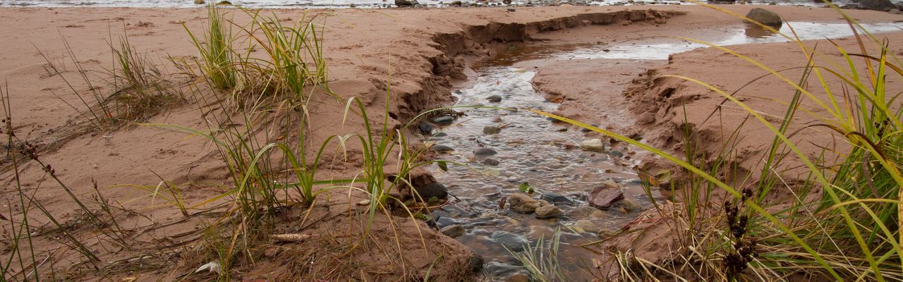 A stream of water runs through a sandy and rocky area and into a body of water in the Keweenaw Peninsula.