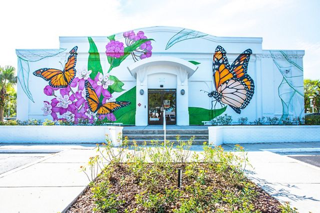 The front of a white building is painted with huge monarch butterflies sitting on plants with green leaves and bright pink flowers. Shrubby plants grow along the front of the building.