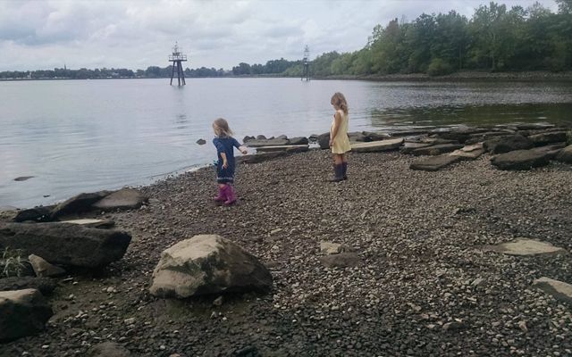 Two young girls wearing rubber boots explore the shoreline of a marsh. One girl has her arm pulled back ready to throw a rock into the water.