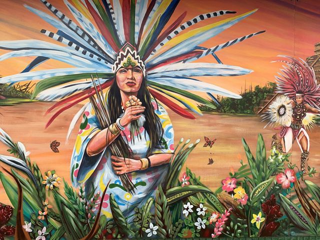 A mural shows two people wearing headgear and clothing decorated with long, colorful feathers, representative of Mexican indigenous cultures. Several painted butterflies fly over flowers.