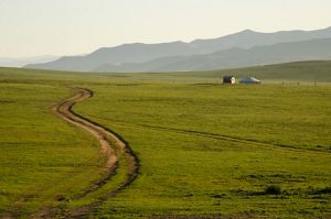 A dirt road winds through open green grassland with a yurt and an outbuilding in the middle ground and mountains in the far distance.