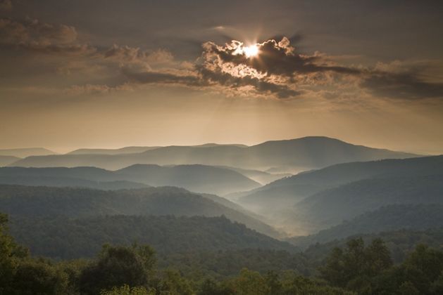 Mountain ridges roll to the horizon in Monongahela National Forest. The sun is partially obscurred by a cloud bank, bathing a deep valley below in soft light.