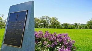 A large granite memorial with an inset brass plaque sits in an open green space. A cluster of pink flowers bloom at the monument's base.