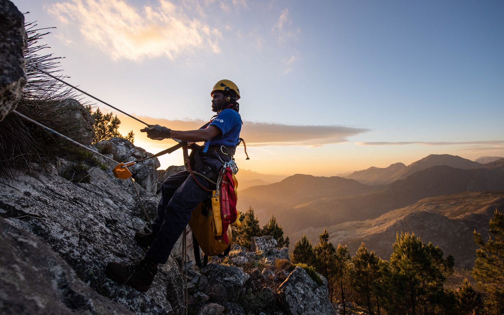 Man climbing down a cliff on a rope against backdrop of a sunrise.