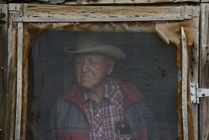 A weathered rancher in plaid stares out his screened window.