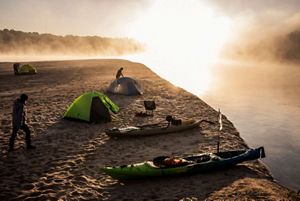 Silhouettes of three campers in fog and early morning light as they move around the campsite along a sandy bank of an island in the river. 