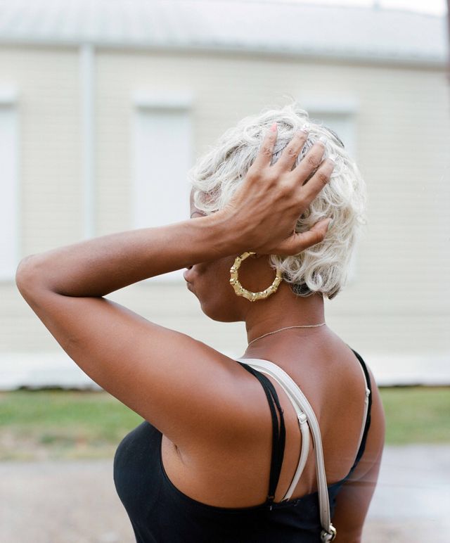 A woman is in the frame from torso up. she is wearing a black tank top and gold hoop earrings. She is in profile but her arm is raised and her hand is touching the back of her head, obscuring her face