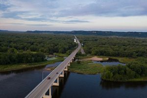 Aerial view of a raised road crossing the Mississippi River near Wabasha, MN. The road leads off into the distance, through dense forested land toward hills in the far distance.