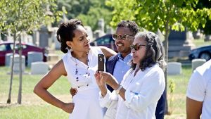 Three people stand together during a family gathering. One of the people uses her cell phone to take a picture of someone out of view.