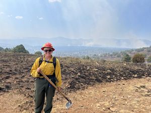 A woman wearing a yellow fire jacket, black pants, and a red helmet holds fire ax while standing outside with mountains in the background. 