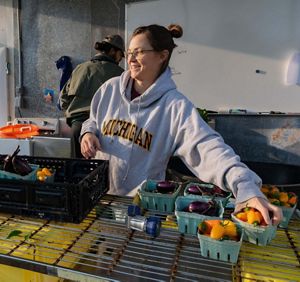 A woman in a grey hoodie smiles while reaching for a small blue container of peppers. A person is working behind her with their back to the camera. 