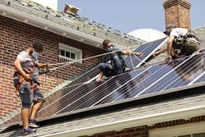 Three workers wearing masks up on a roof installing solar panels. Hilario Minaya is on the left, standing on the roof with a harness and rope for safety.
