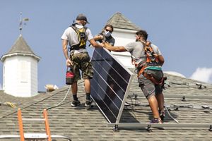 Three masked workers, viewed at center, on top of a roof installing a solar panel.