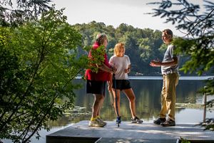 Three people stand on a dock by a lake, talking and gesturing toward the water.