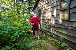 Person with a walking stick walks along a wooded path beside a rustic cabin.