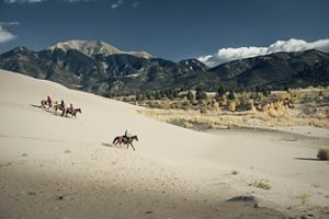 Horseback riders ride across a sand dune at the base of mountains.