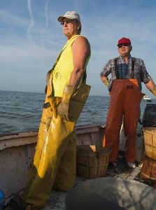 Two watermen at work on a skiff. Only the lower half of the men are visible. They're wearing yellow and orange waders. Empty bushel baskets are at their feet.