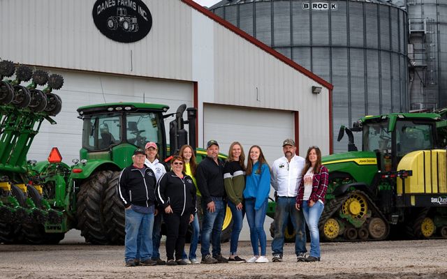Nine members of the Danes family stand and pose in front of green tractors and metal buildings of Danes Farm.