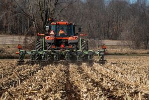 A red tractor with green blades drives away from the camera as it works in a golden field of cut cornstalks.
