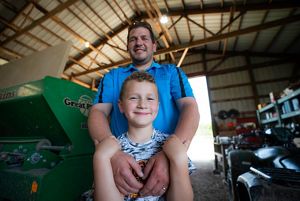 Portrait of Ryan Schulz with his arms around his son Dylan, who stands in front of him, inside a farm building.