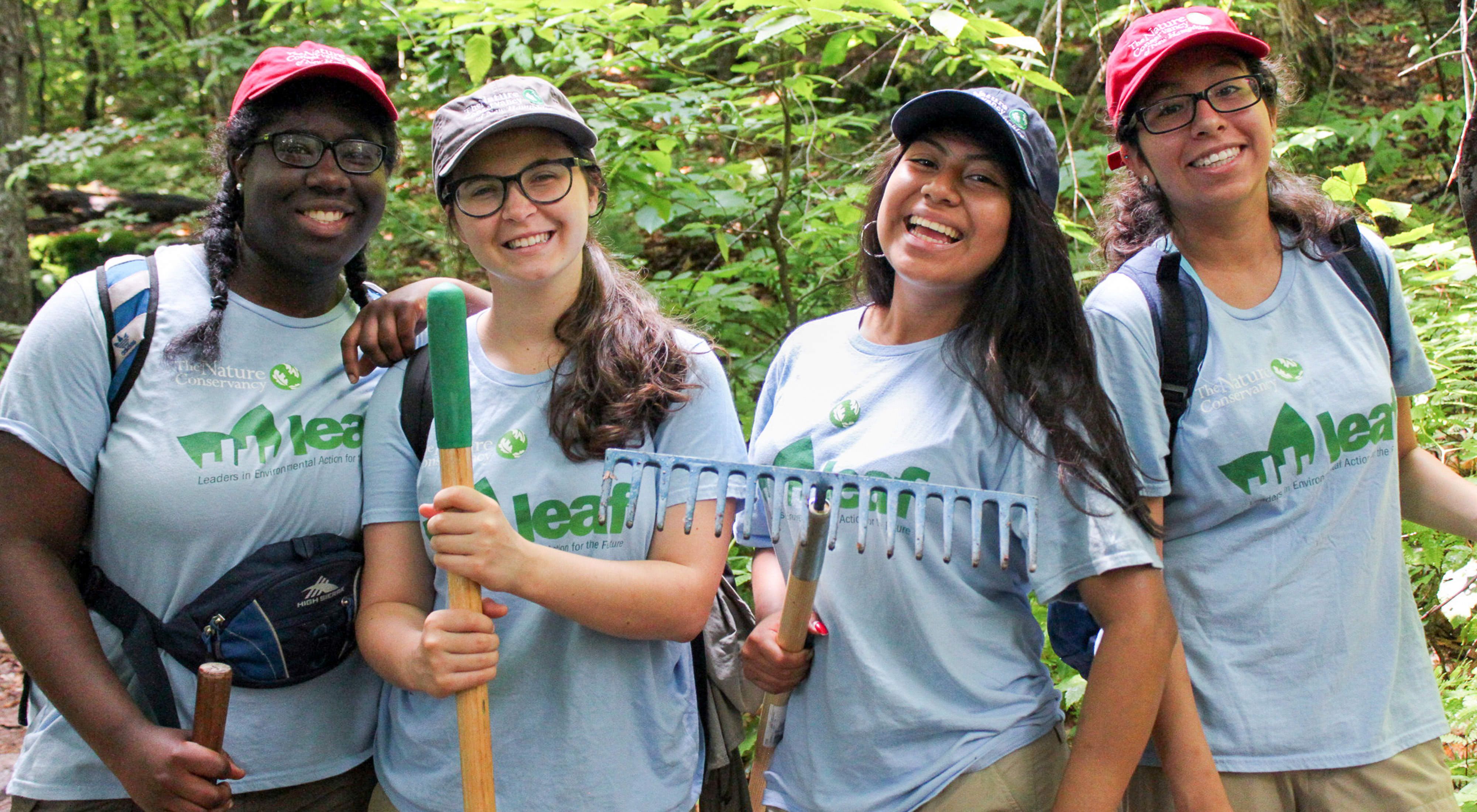 Four young women in blue t-shirts smile at the camera while holding rakes and shovels.