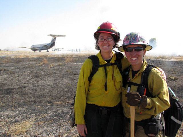 Two women pose together during a controlled burn. Their smiling faces are streaked with soot.