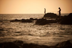 Two people fishing on the rocks of a shoreline at dusk.