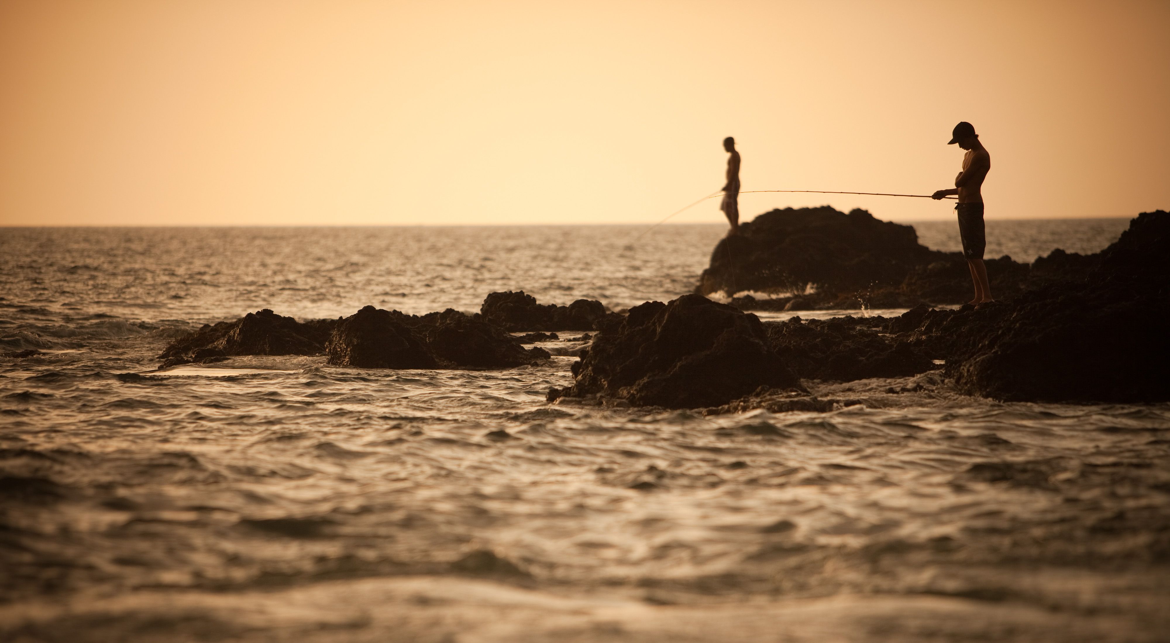 Two people fishing on the rocks of a shoreline at dusk.