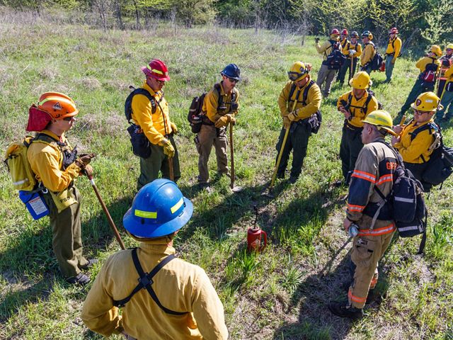 Nearly fifteen fire practioners in fire gear stand in a circle on the green grass of Matthews Prairie during a briefing before a prescribed fire. 